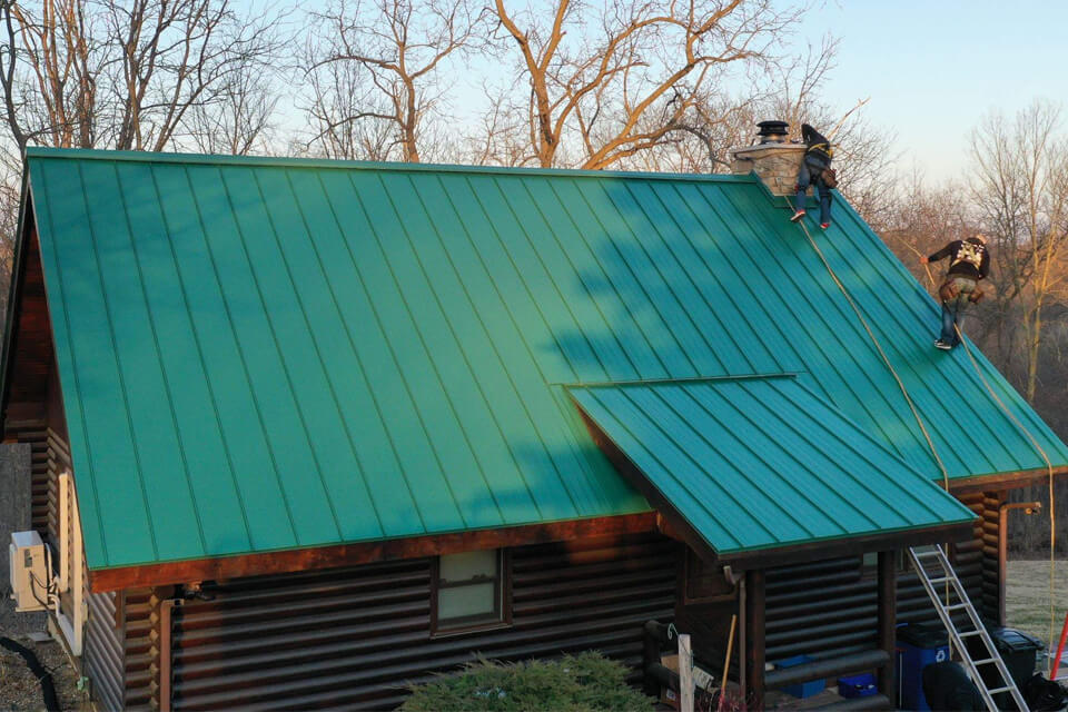 Photo of a log cabin and 2 roofers installing a green metal roof