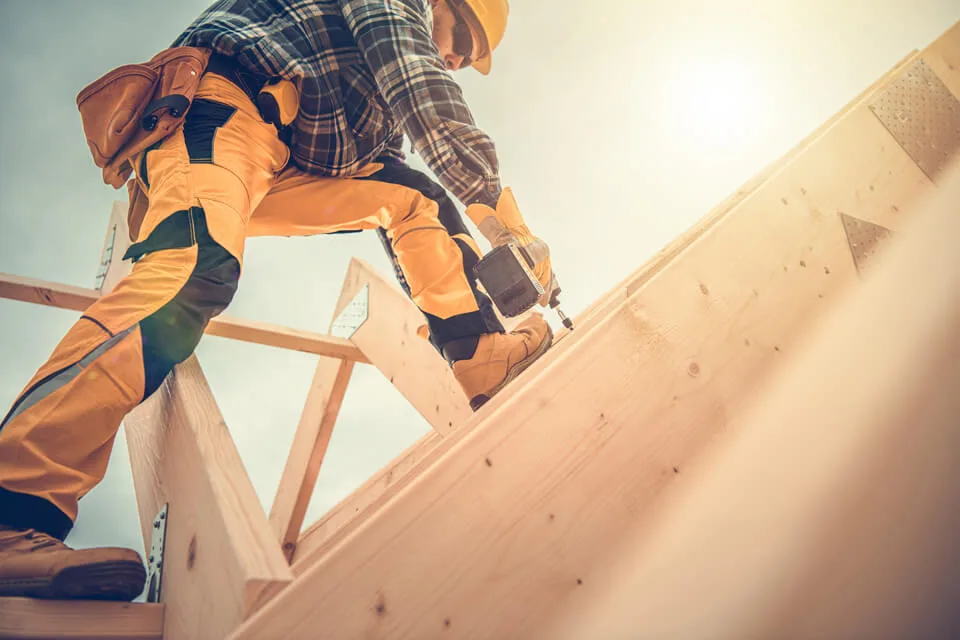 Roofer in a plaid shirt and orange work pants standing on the frame of a roof drilling a screw into a rafter