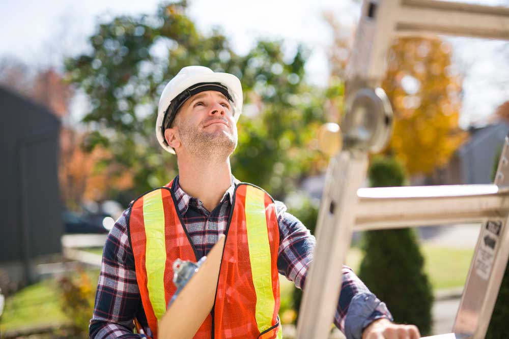 Roof inspector with a hard hat and orange vest holding a ladder and looking up